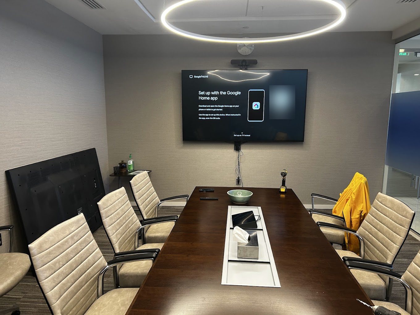 A 75 inch TV with a camera above it, mounted at the end of a long conference table.