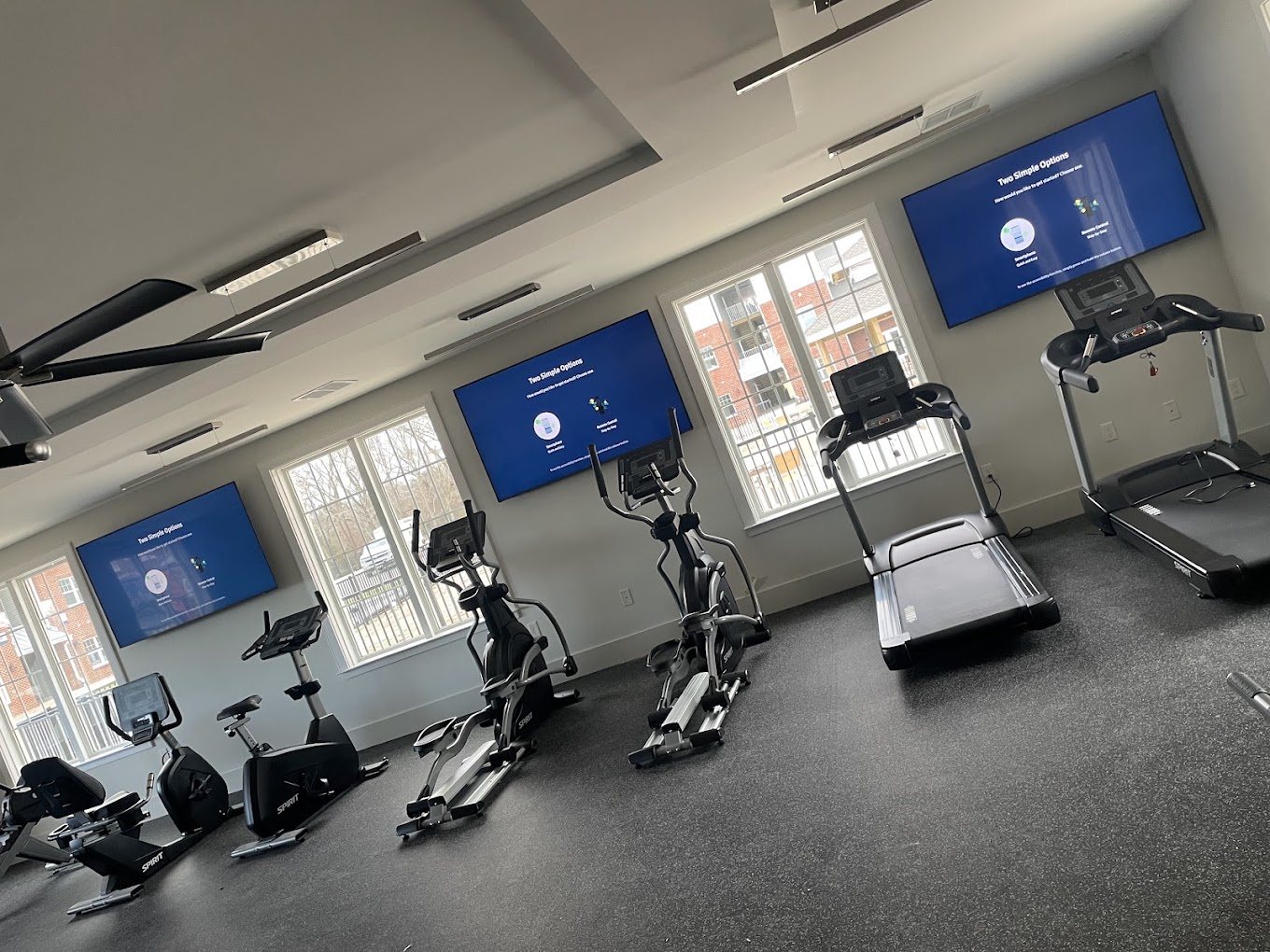 Three 65 inch TVs mounted high on the wall above treadmills and ellipticals in a fitness center.
