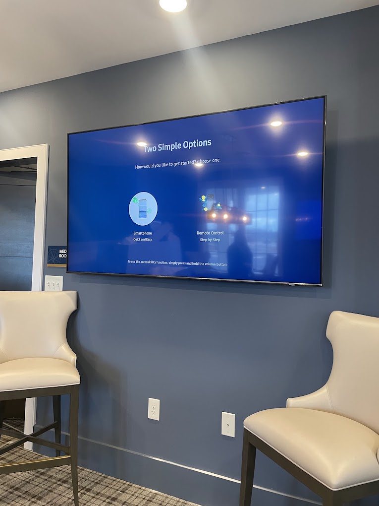 A 75 inch TV mounted on a blue wall in an office lounge with cream chairs.