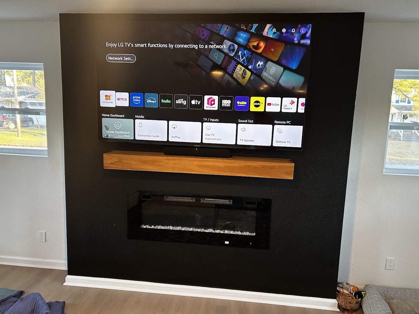 A 75 inch TV mounted on a black feature wall above a floating wood shelf and an electric fireplace.